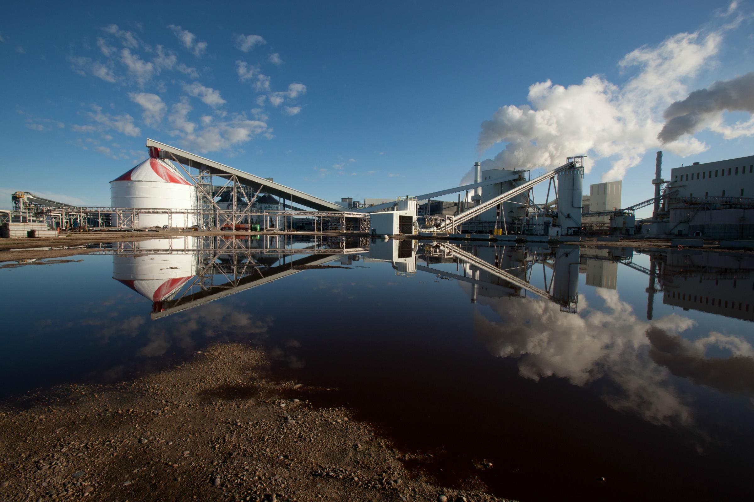An industrial facility for mining with conveyors and storage buildings with a large puddle of water in the foreground reflecting the scene
