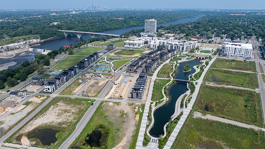 Aerial image of an urban, residential development site next to a large river with a long, narrow pond extending through the middle on a sunny day.