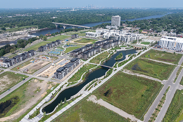 Aerial image of an urban, residential development site next to a large river with a long, narrow pond extending through the middle on a sunny day.