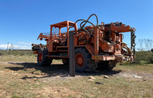 Drilling rig conducting geotechnical testing in desert terrain near power-utility infrastructure.