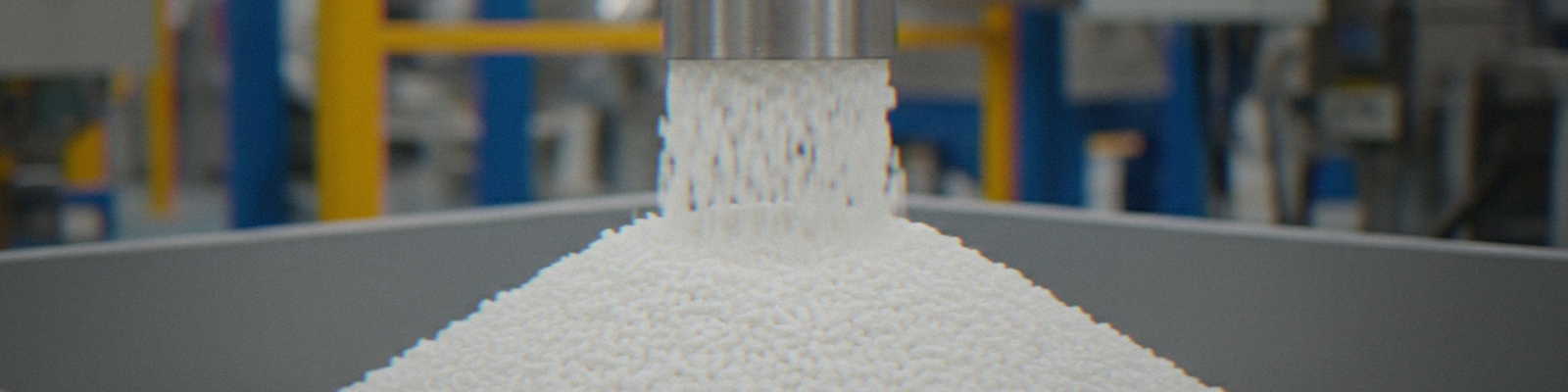 White plastic polymer pellets pour out from an industrial machine into a large storage container in a manufacturing facility.