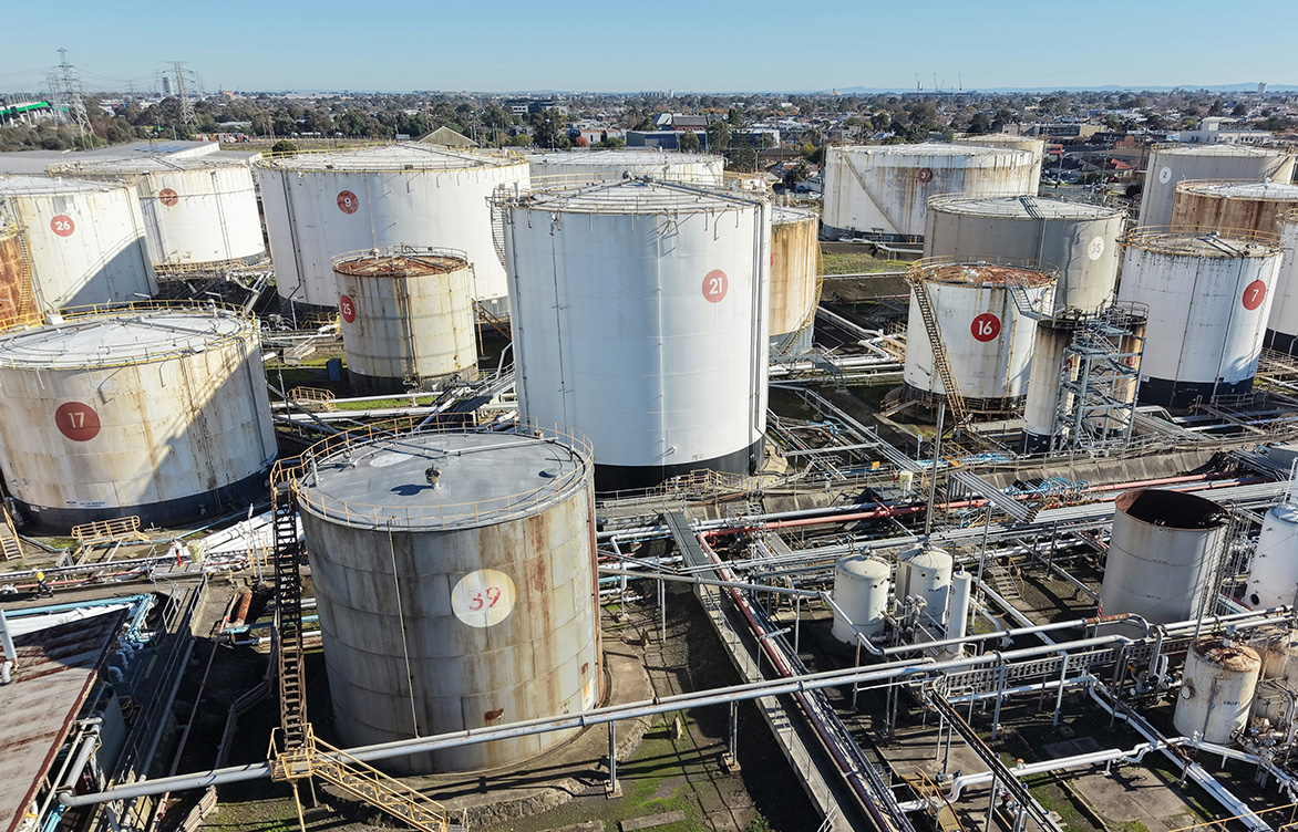 Industrial fuel storage facility with multiple large cylindrical tanks, interconnected pipelines, and processing equipment viewed from above.