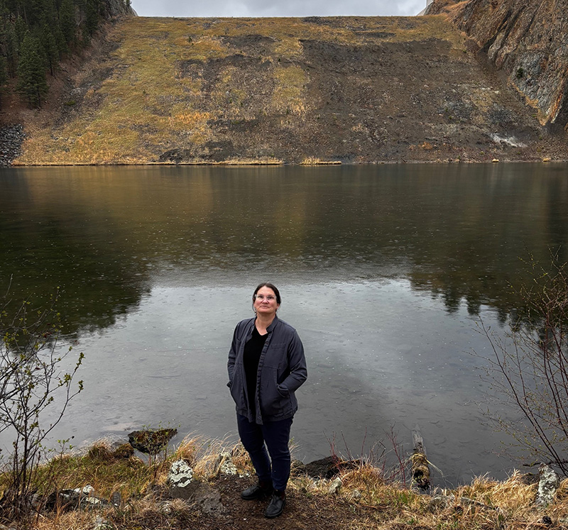 An engineer in field clothing stands in drizzling rain in front of an earthen reservoir spillway