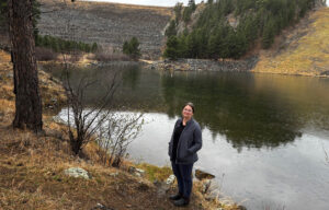 An engineer in field clothing stands in drizzling rain in front of an earthen reservoir spillway.