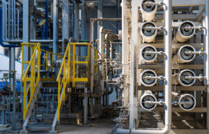 A wide shot of a complex industrial water treatment facility. Multiple white reverse osmosis membrane pressure vessels are arranged in a rack, connected by a network of stainless steel pipes and valves, and by a metal staircase with yellow safety handrails.