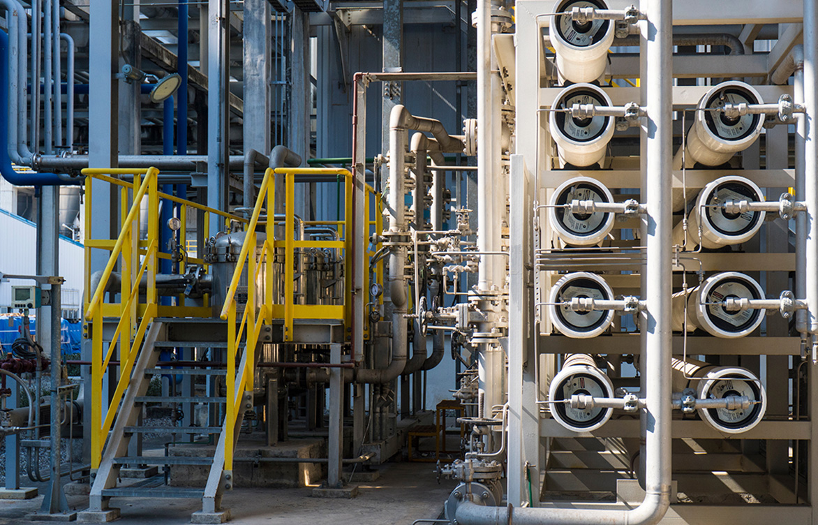 A wide shot of a complex industrial water treatment facility. Multiple white reverse osmosis membrane pressure vessels are arranged in a rack, connected by a network of stainless steel pipes and valves, and by a metal staircase with yellow safety handrails.