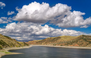 Reservoir surrounded by dry, rolling hills under dramatic white clouds and a bright blue sky.
