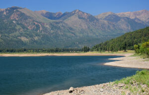 Reservoir surrounded by mountains, conifer forests, and a cloudless sky.