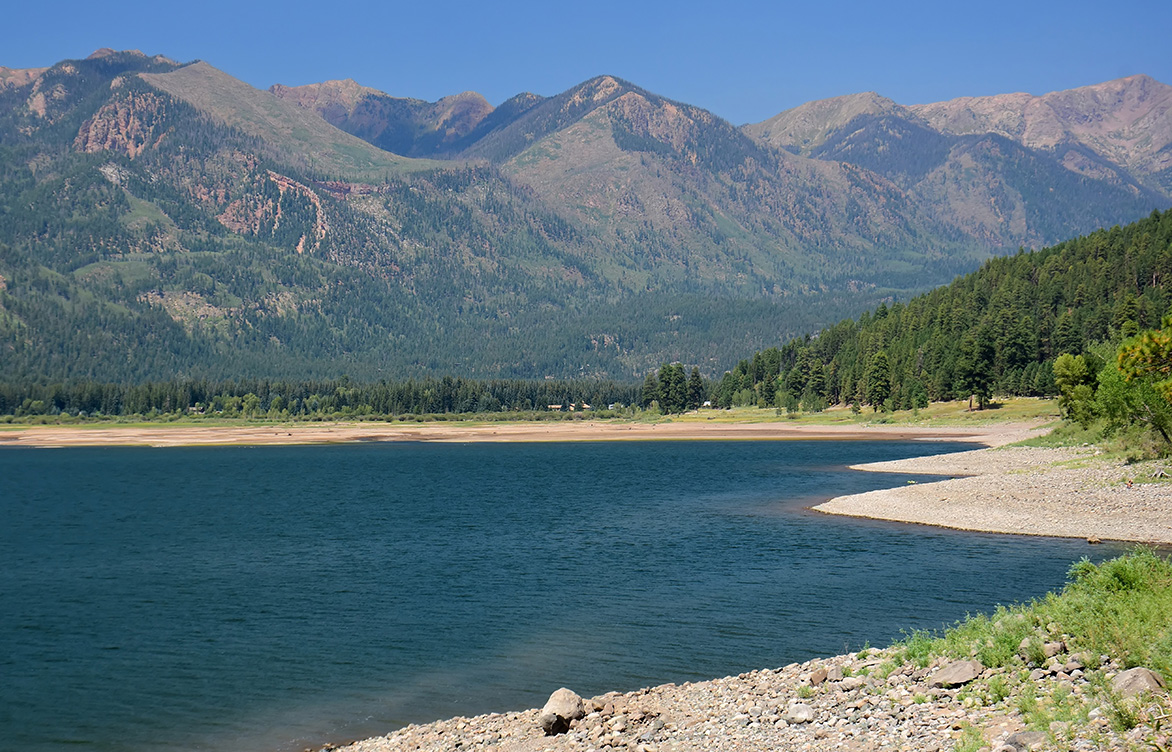 Reservoir surrounded by mountains, conifer forests, and a cloudless sky.