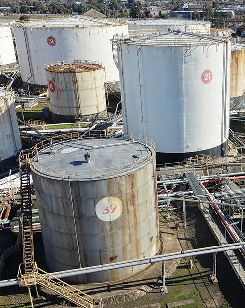 Industrial fuel storage facility with multiple large cylindrical tanks, interconnected pipelines, and processing equipment viewed from above.