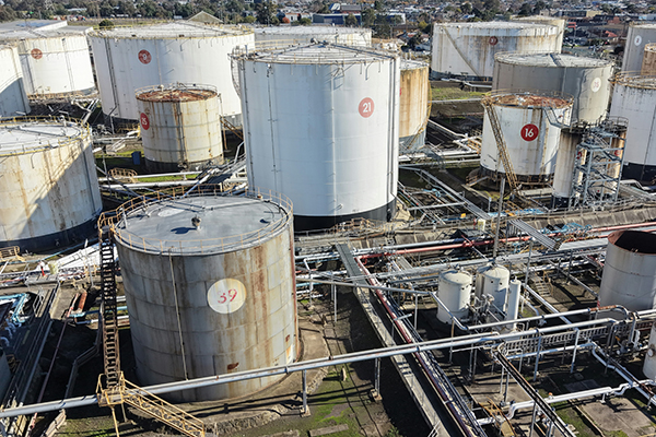Industrial fuel storage facility with multiple large cylindrical tanks, interconnected pipelines, and processing equipment viewed from above.