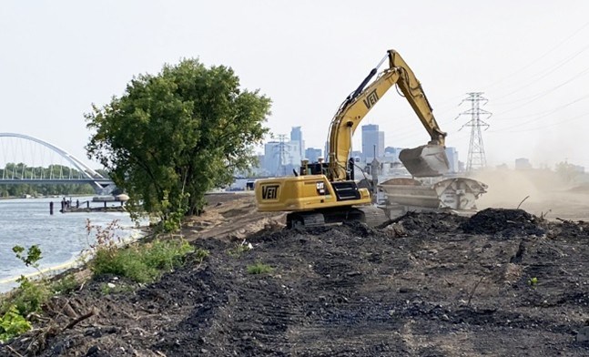 Excavator placing material along a riverfront construction site, with a bridge and city skyline visible in the background.
