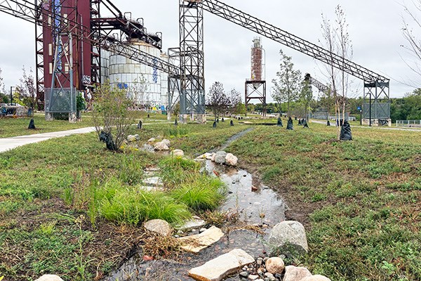 Stormwater channel with native vegetation and rocks running through a restored industrial site beneath overhead conveyor structures.