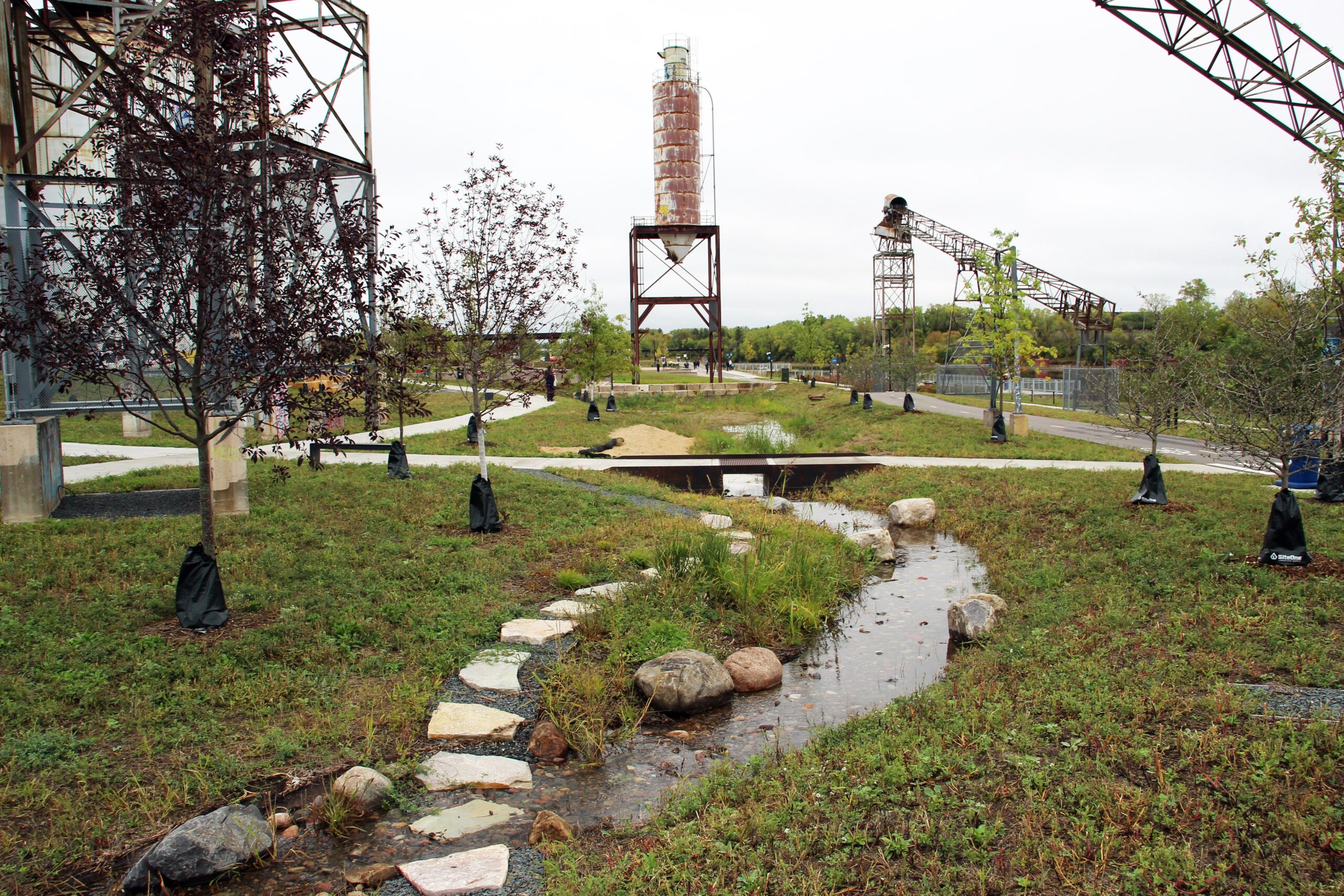 Meandering stormwater channel with rocks, native vegetation, and young trees integrated into a restored riverfront landscape alongside preserved industrial structures.