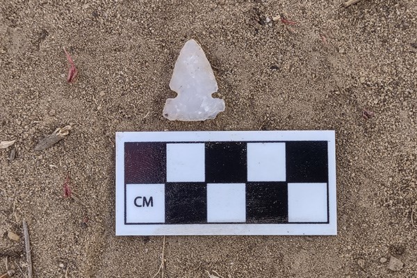 A triangular, notched arrowhead of white stone lies on sand next to a black-and-white scale card.
