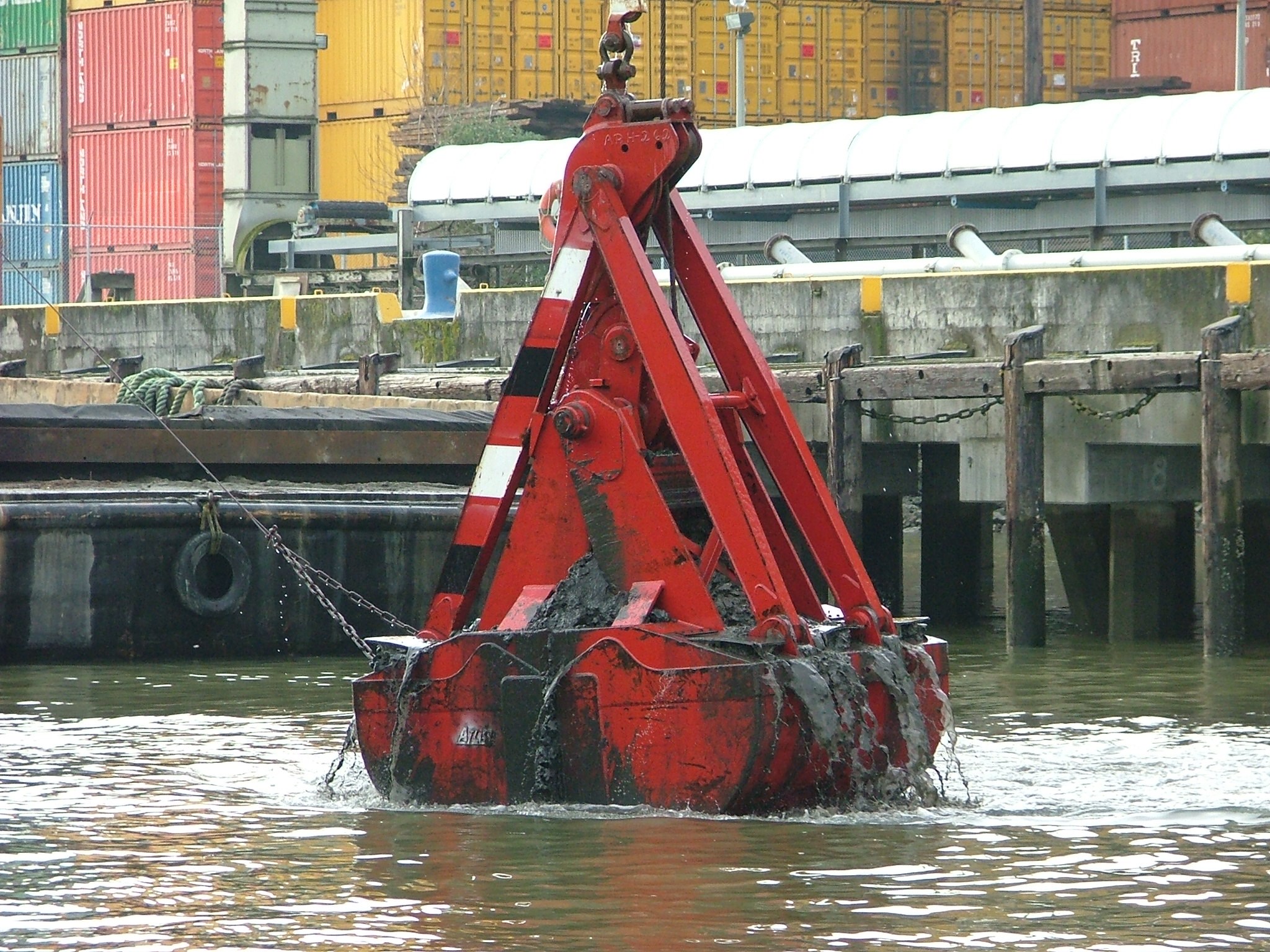 Industrial clamshell dredge bucket lifting sediment from water at a dock with stacked shipping containers in the background.