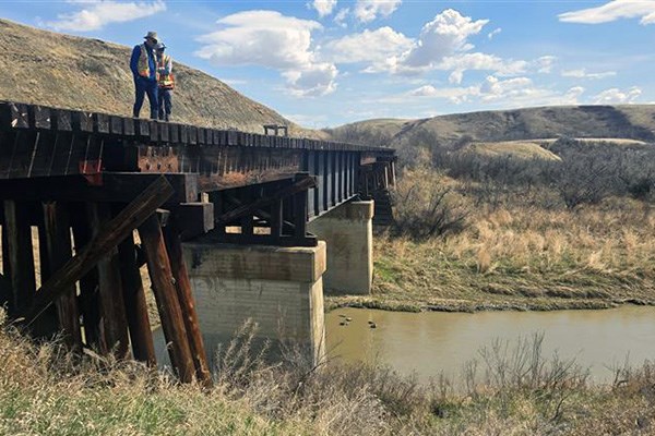 Two workers in safety gear stand on an elevated wooden trestle bridge supported by concrete piers, overlooking a narrow river in a grassy, hilly landscape under a partly cloudy sky.