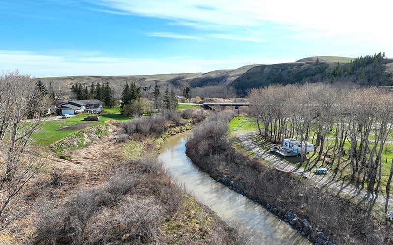 A view of a sunny river valley, looking downstream, with sparse buildings on either bank and mountains in the background.