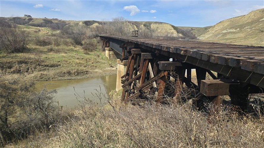 Old wooden railroad trestle bridge spanning a shallow river in a dry, grassy valley with rolling hills.